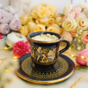 Gros plan d’une bougie dans une tasse bleu profond ornée de motifs grecs dorés.